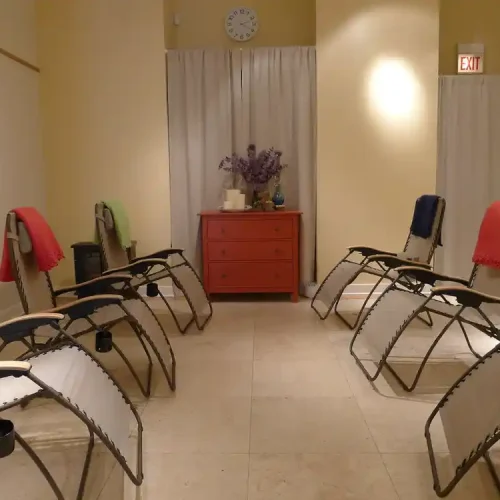 A quiet room with eight reclining chairs arranged in a circle, each draped with a blanket, and a red dresser with a flower arrangement at the far end. healing found