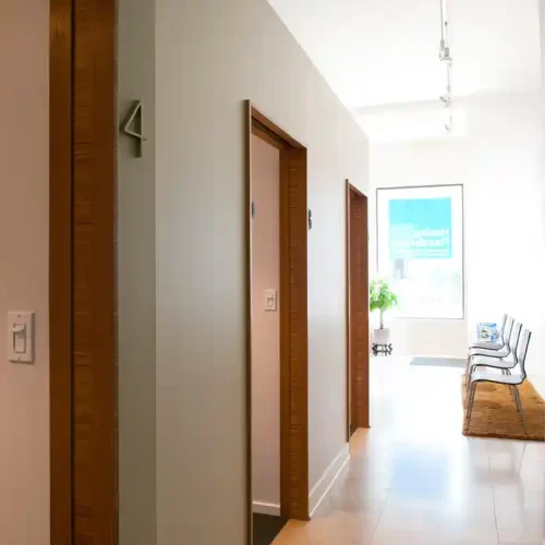 Clean hallway with numbered doors, light-colored flooring, a plant near the window, and chairs along one side. Healing found
