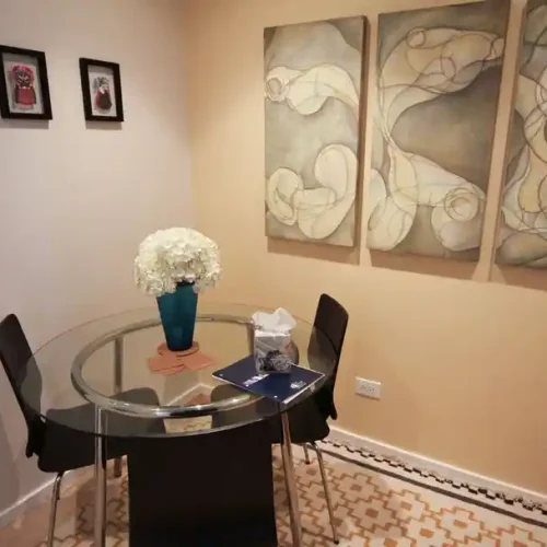 Small dining area with a glass table, black chairs, a vase of white flowers, and abstract artwork on the wall.