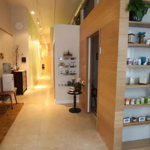 Modern hallway with shelves displaying small items, a water cooler, chairs, and artwork on the walls. A door leads to another room at the end.