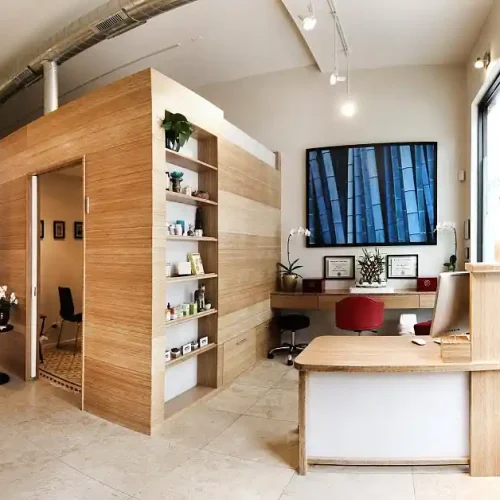 Modern office space with wooden shelving, a desk with a computer, and a plant-filled corner. A wall partition separates a small treatment room, and a large window lets in natural light.Healing found