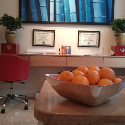 A modern office with a bowl of oranges in the foreground, red chairs, framed certificates on the wall, and potted plants on a desk healing found