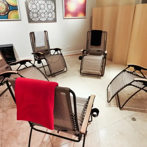 A relaxation room with five reclining chairs, each with a towel draped over them. Colorful artwork hangs on the walls, and wooden partitions separate the space.
