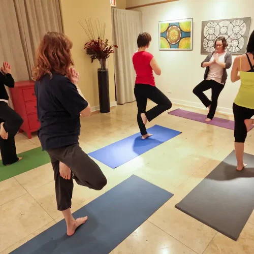 A group of five people practicing yoga in a studio, each on their own mat, performing a tree pose. Colorful artwork hangs on the wall, and a heater is visible in the background. Healing found