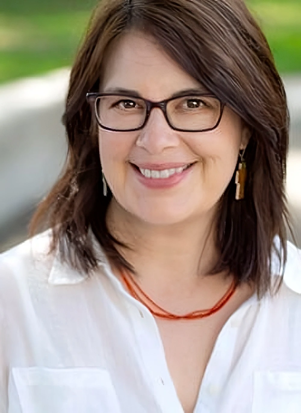 A smiling woman with glasses and dark brown hair, wearing a white blouse and orange necklace healing found